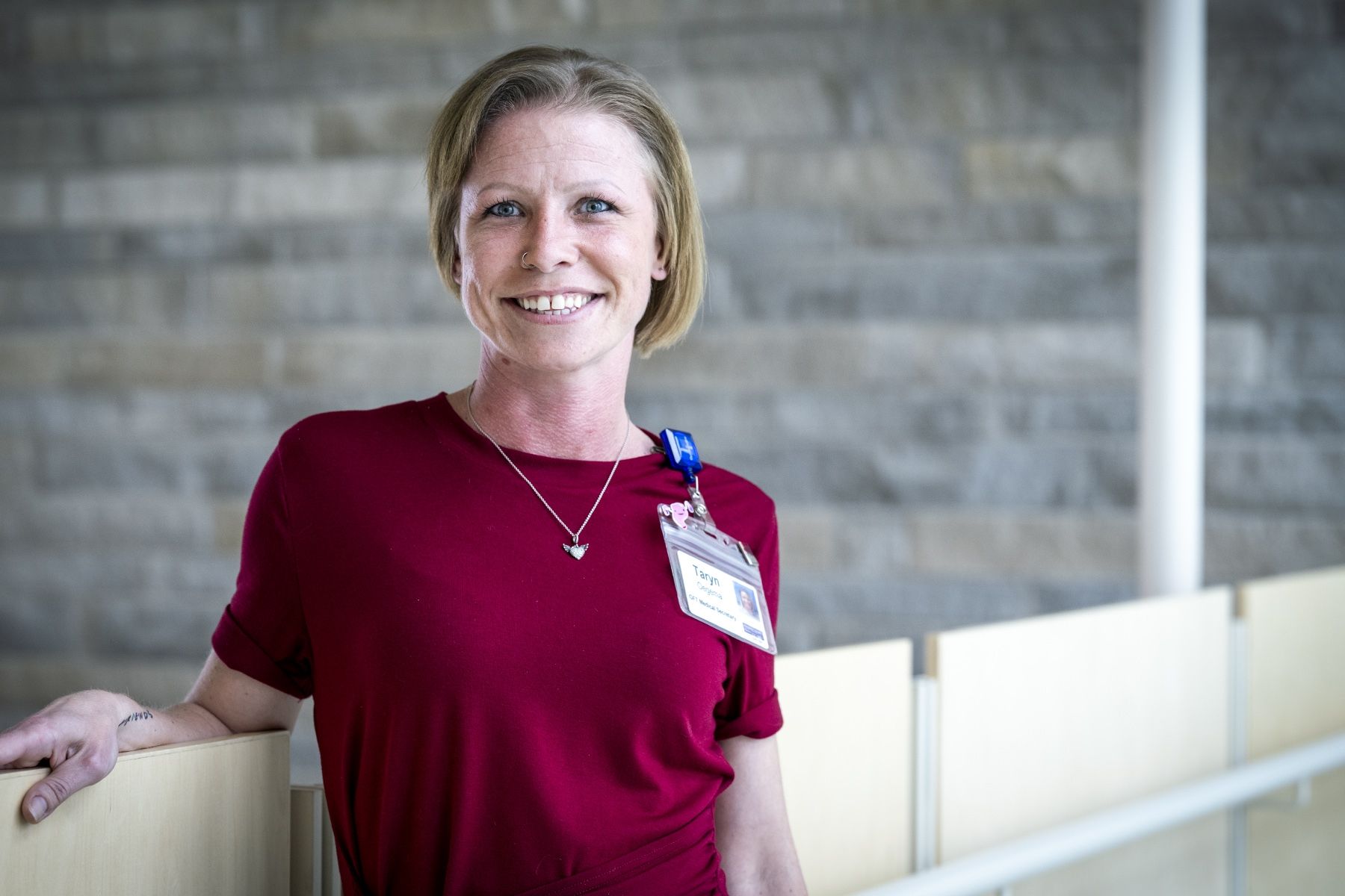 Taryn Oegema is pictured standing and leaning against a bannister in a hallway at Kingston General Hospital. She has short blonde hair, blue eyes and has a big smile on her face. She’s wearing a dark red blouse. 