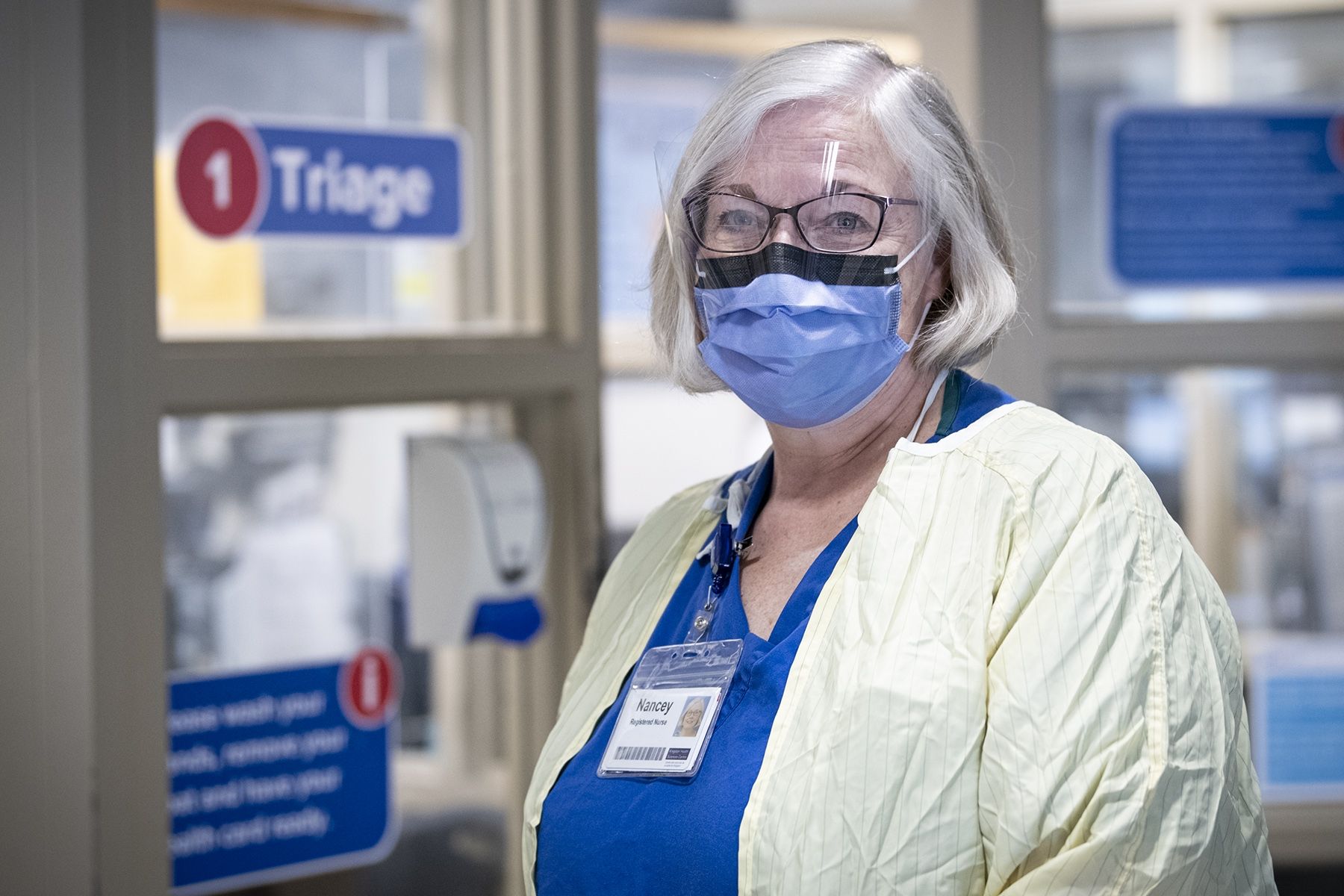 Nancey Martindale pictured inside KHSC's Emergency Department has short grey hair. She wear glasses and is photographed wearing a mask, blue scrubs and a yellow gown.