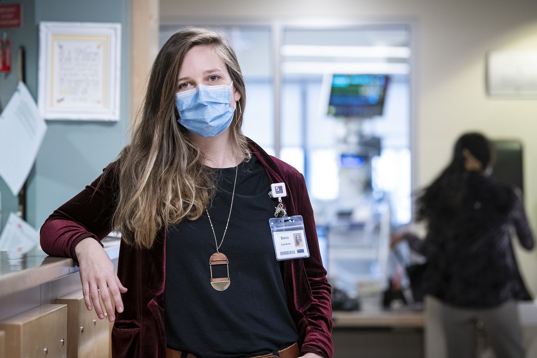Bianca Sabatini is pictured leaning on the care desk at the Intensive Care Unit (ICU) at the KGH site. She has blue eyes and medium brown, wavy hair that goes down past her shoulders. She’s wearing a black top with a burgundy blazer on top. 