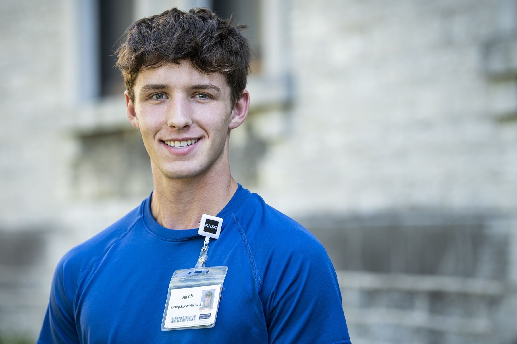 Jacob Kasaboski has short, dark, curly hair. He's wearing a bright, blue top and standing outside the Kingston General Hospital site.