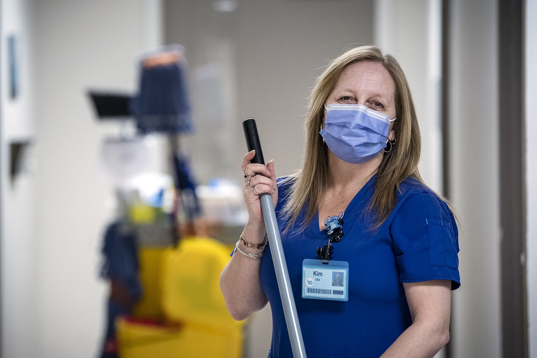 Kimberley Dickinson is pictured holding a broom in a hallway at the HDH site. She has blonde, shoulder length hair and is wearing blue scrubs and mask.