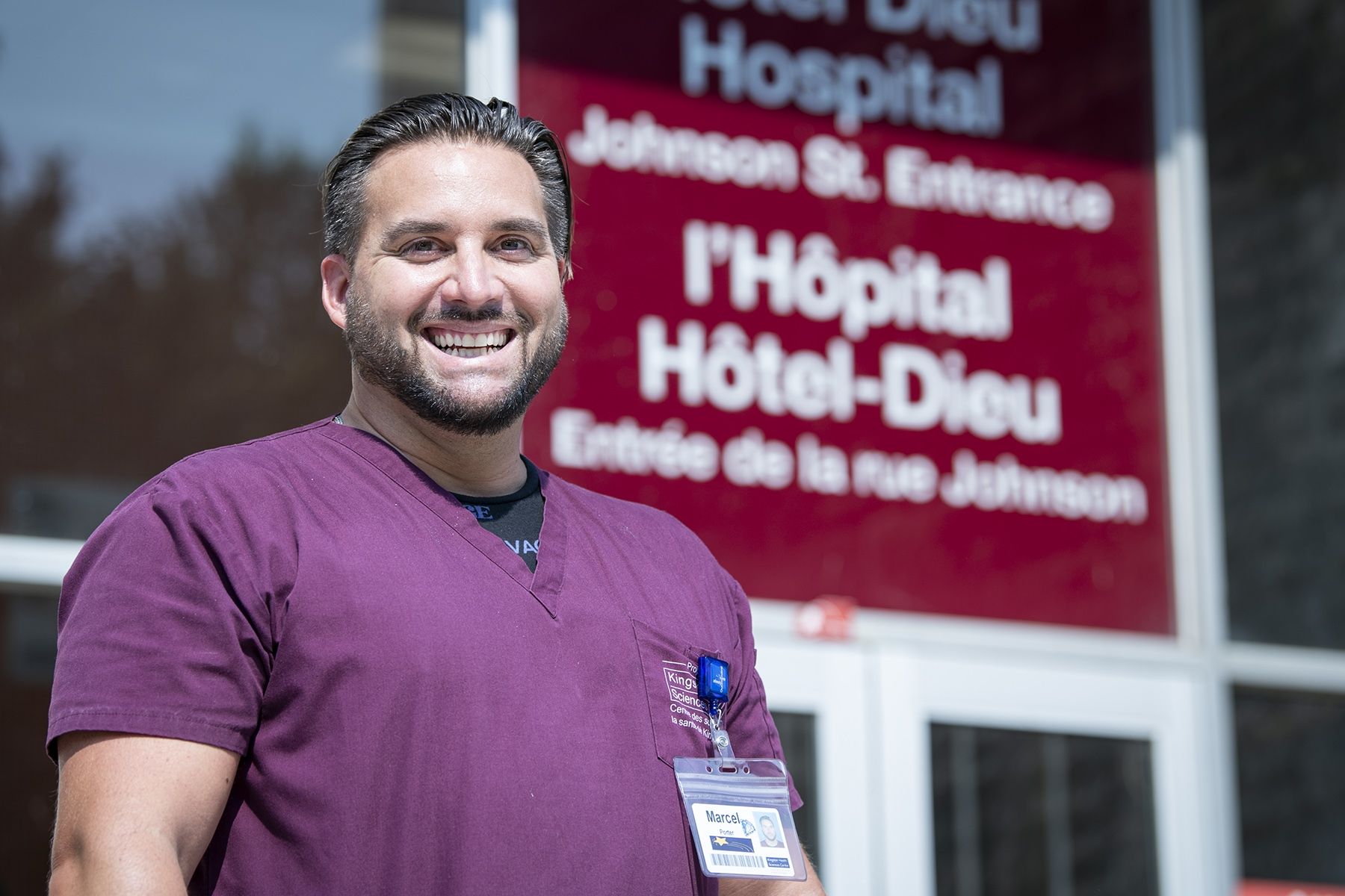 Marcel Cernik has short dark hair and a beard. He's wearing purple scrubs and pictured standing outside of Hotel Dieu Hospital.