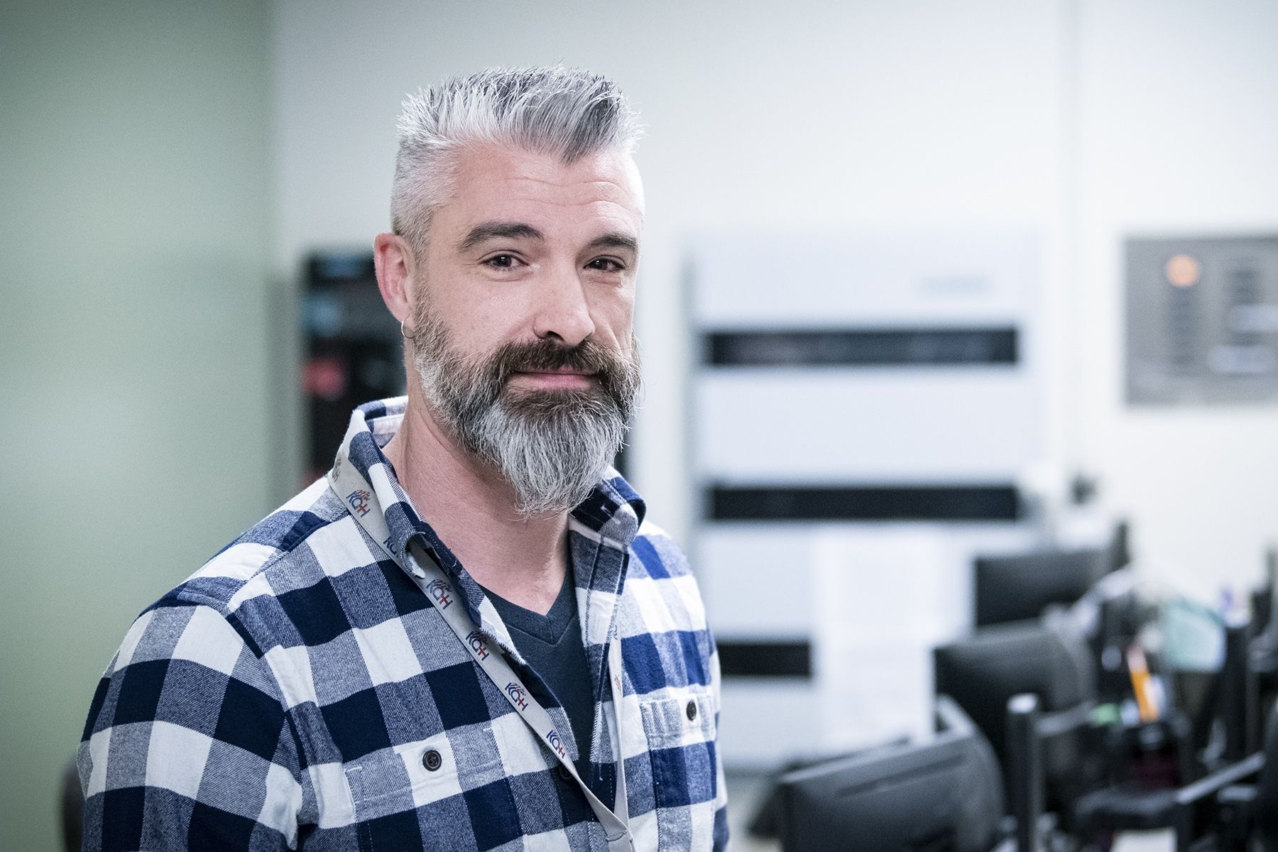 Mark Westcott is pictured in the Switchboard department at the KGH site. He has dark brown eyes, salt and pepper, spiky hair and a beard. He’s wearing a blue and white plaid, long-sleeved shirt. 