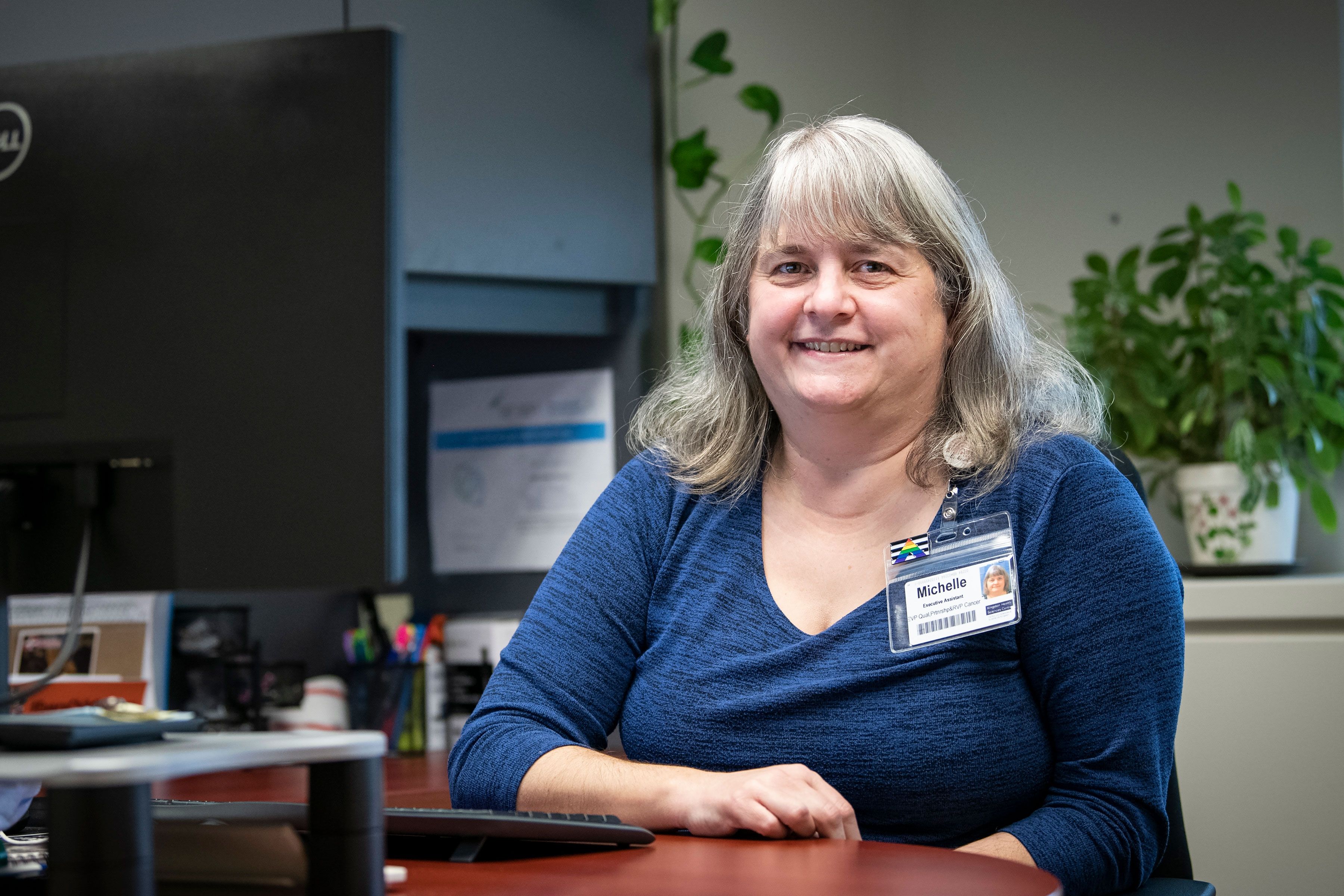 Michelle Hiebert is pictured sitting at her desk, located at the HDH site. She has shoulder-length, silver hair and is wearing a blue, long-sleeved shirt. 