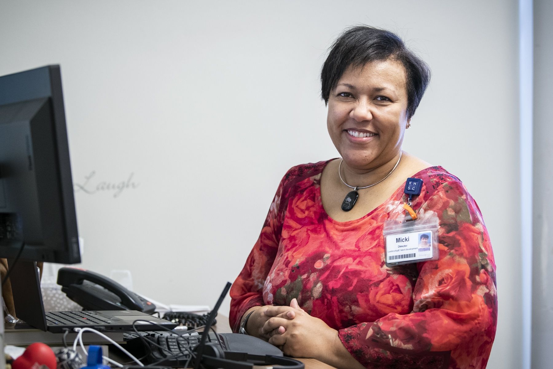 Micki Mulima is pictured sitting at her desk at the KGH site. She has short, dark hair and is wearing a red, patterned top. 